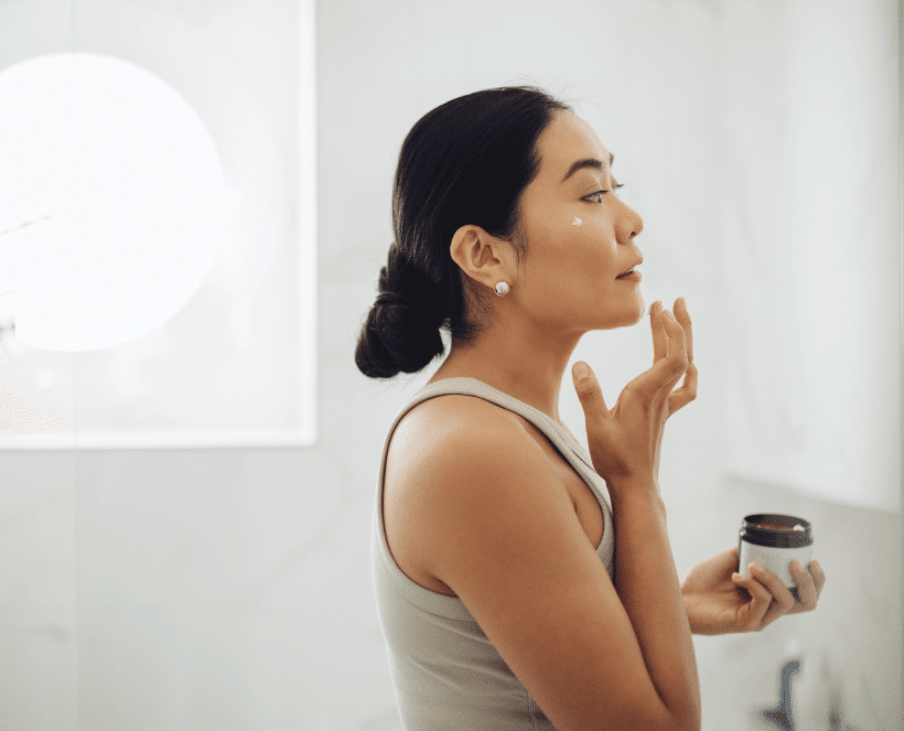 A woman in a light tank top applies cream to her cheek from a small jar in her left hand. Her dark hair is in a bun, and she has a pearl earring.