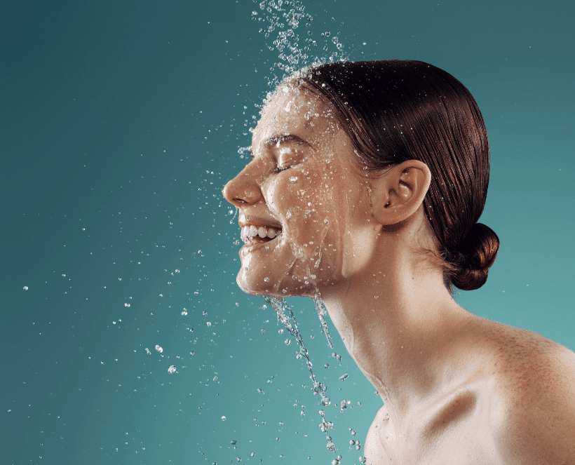 A profile photo of a smiling woman with closed eyes, water splashing on her face and dark, wet hair pulled back in a bun, showing her freckles against a solid teal background.