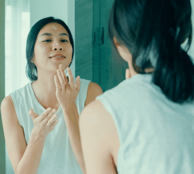 A woman in a light-colored sleeveless top applying white cream to her face while looking at her reflection in a mirror.